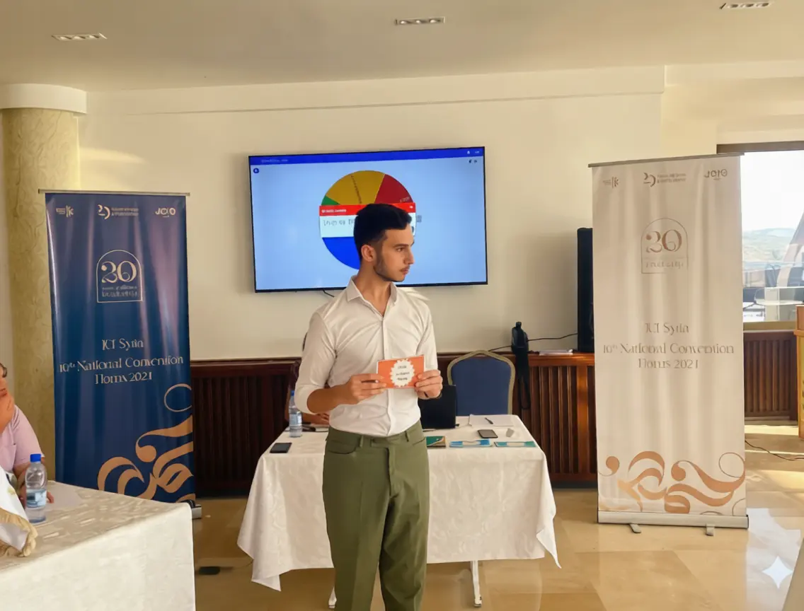 A young man in a white shirt and olive trousers stands in a conference room holding a small card. Behind him is a digital screen showing a colorful wheel and two large banners for the JCI Syria 10th National Convention held in Homs, 2021.